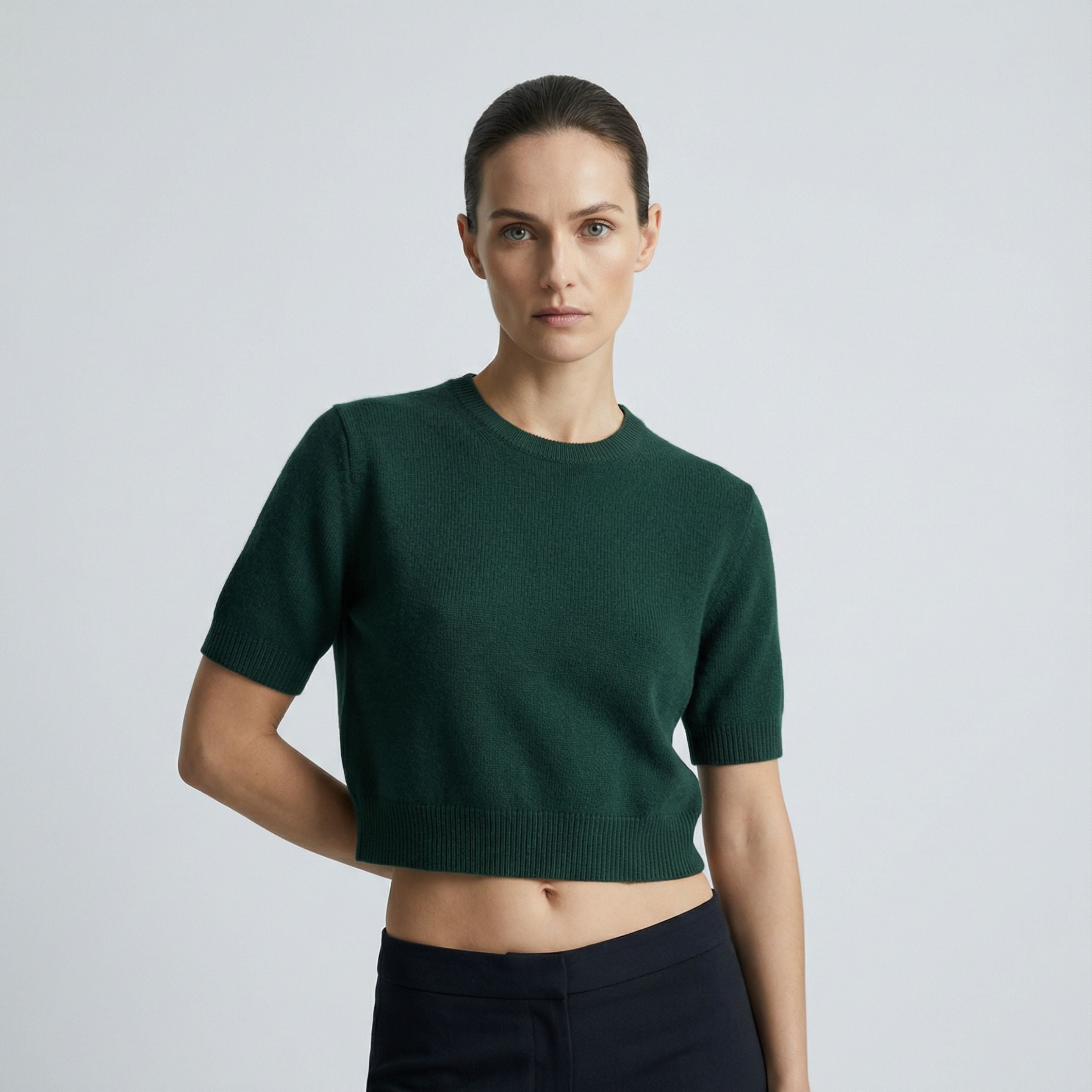Female model wearing a royal green, short sleeve knitted crop top made of 100% cashmere pashmina, paired with dark grey trousers.