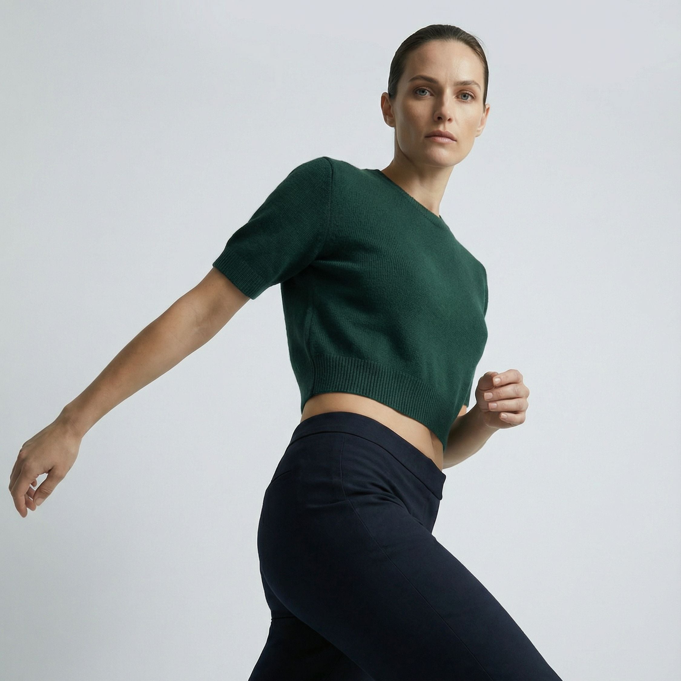 Female model wearing a royal green, short sleeve knitted crop top made of 100% cashmere pashmina, paired with dark grey trousers.