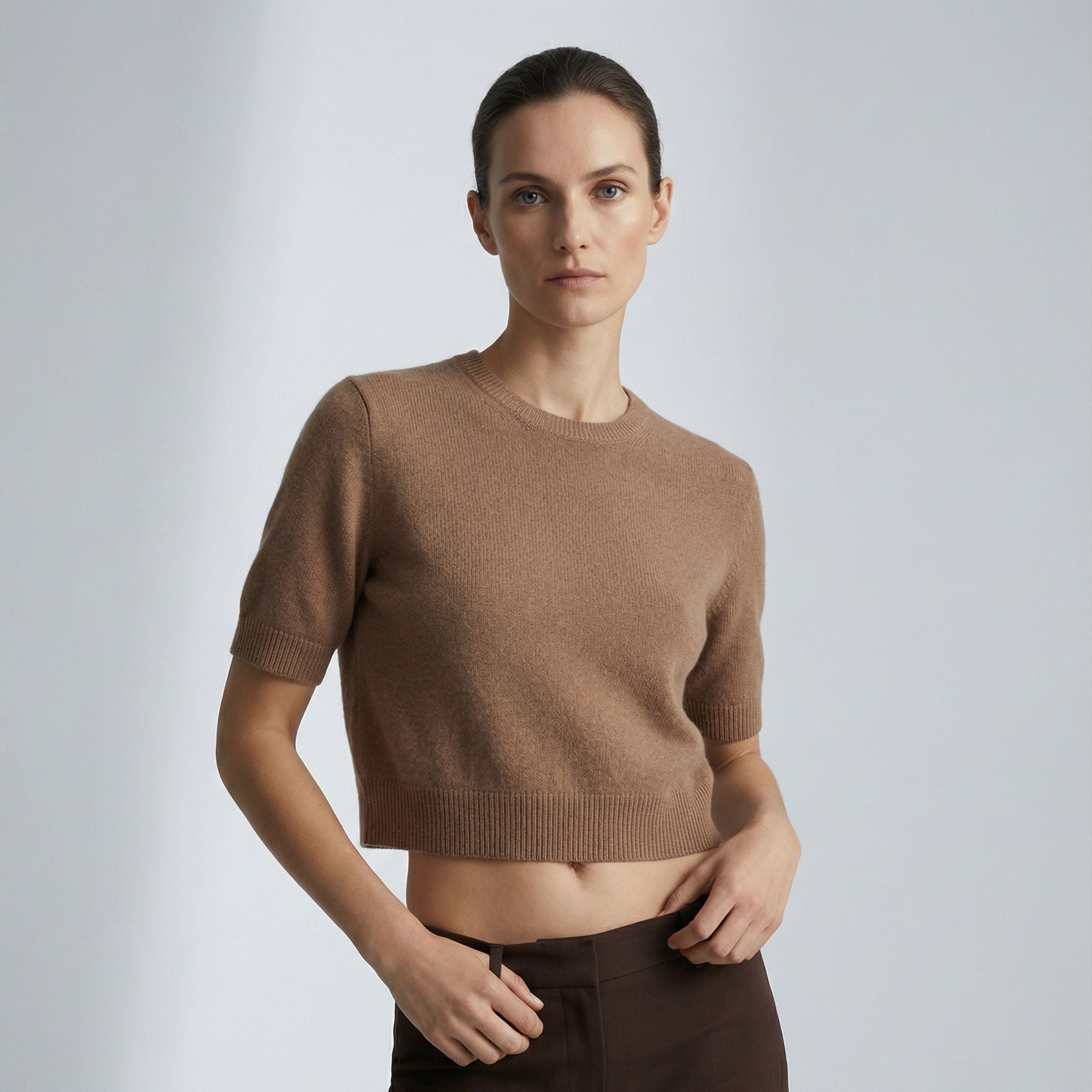 Female model wearing a Beige, short  sleeve knitted crop top made of 100% cashmere pashmina, paired with dark brown trousers.