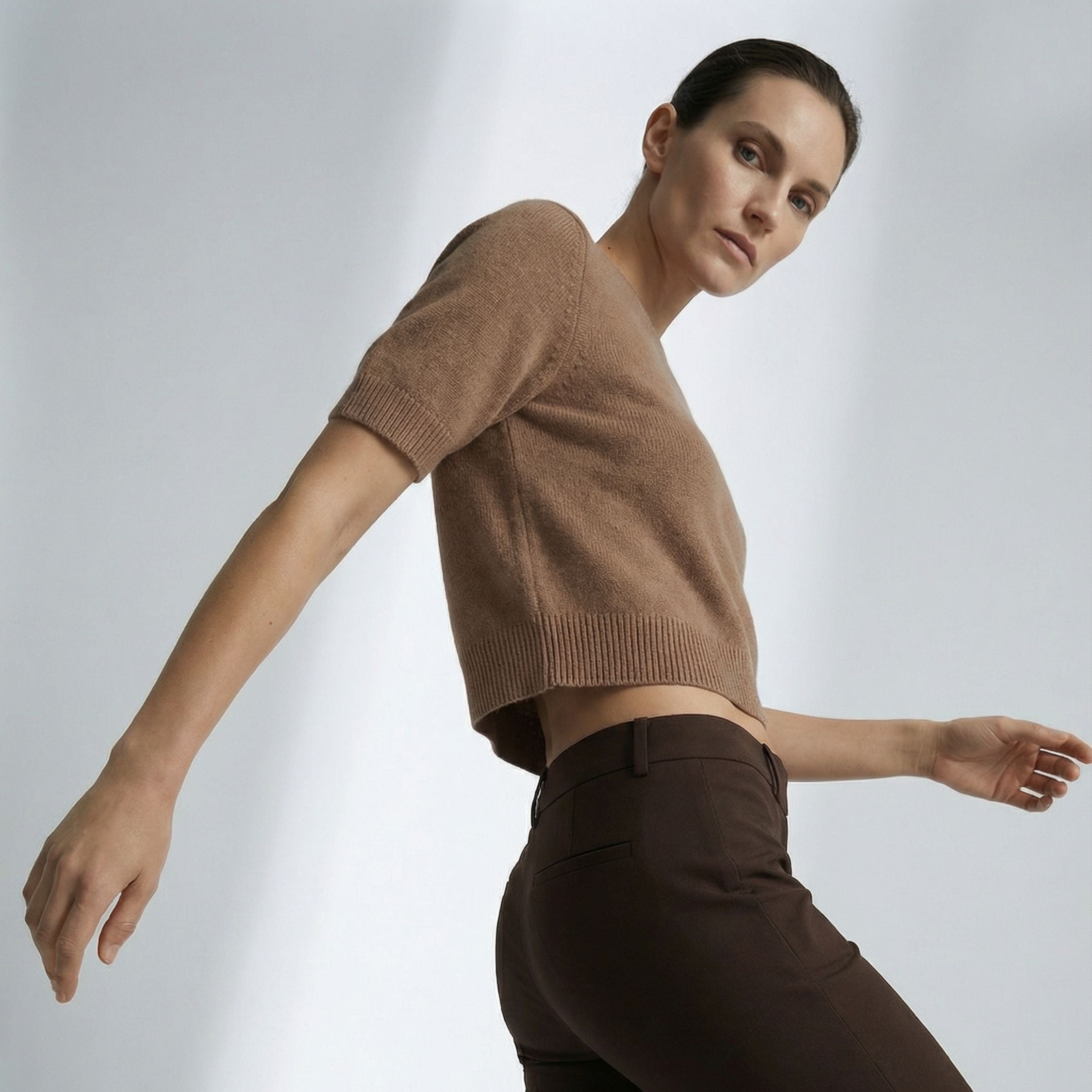 Female model wearing a Beige, short  sleeve knitted crop top made of 100% cashmere pashmina, paired with dark brown trousers.