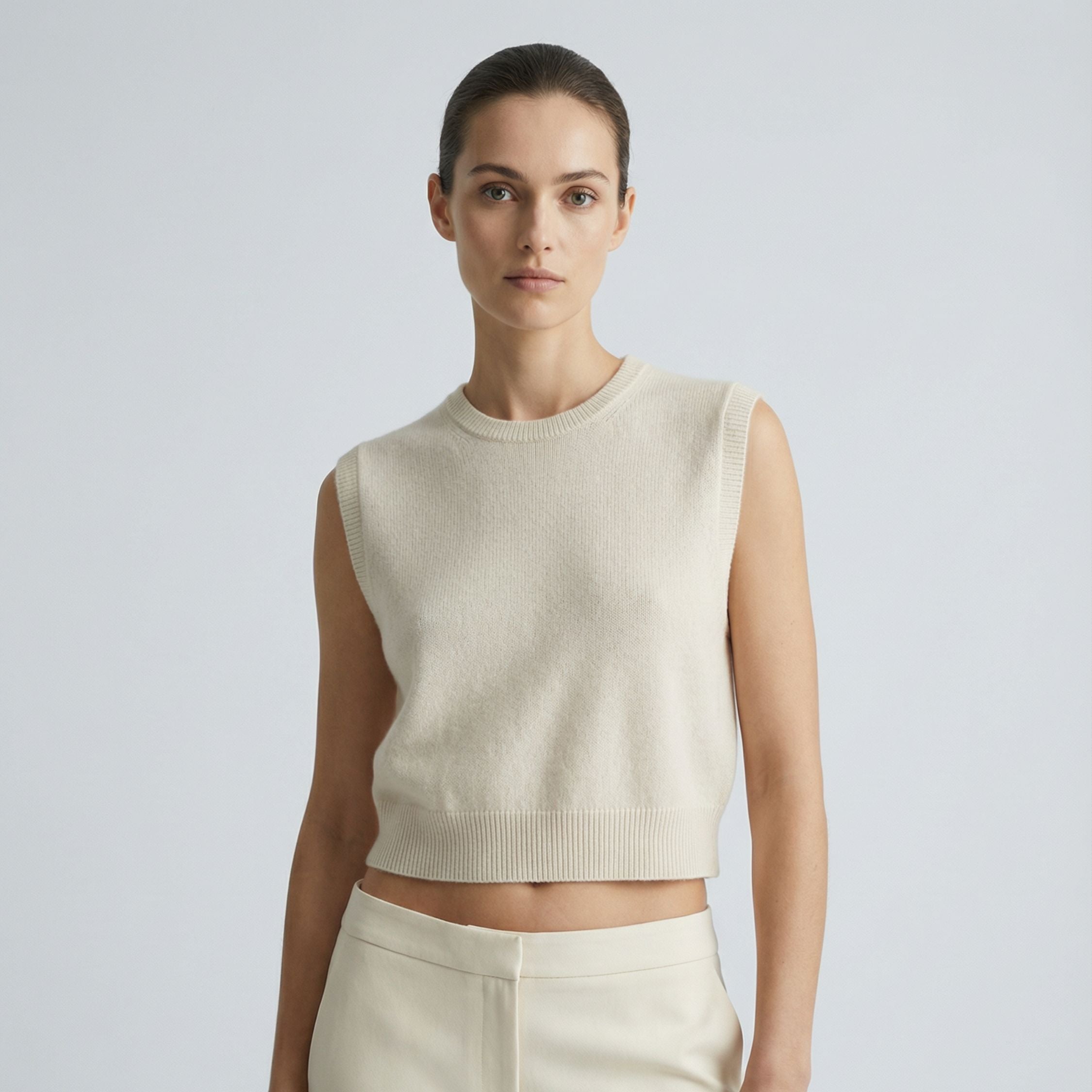 Female model wearing a cream white, sleeveless knitted crop top made of 100% cashmere pashmina, paired with cream white trousers.